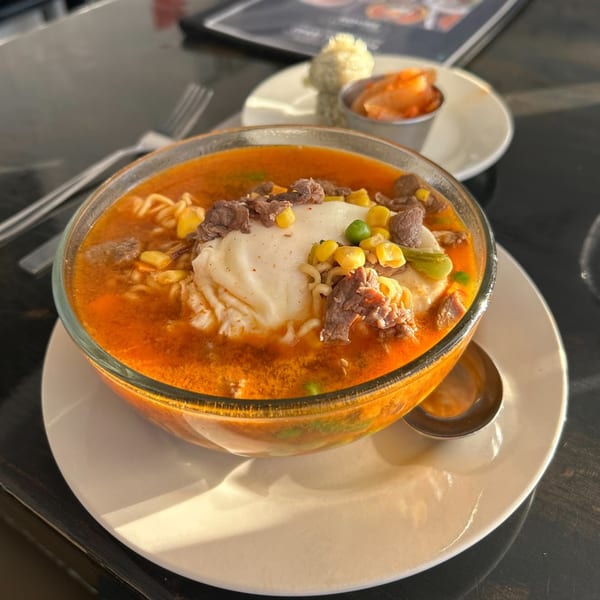 A bowl of spicy ramen with beef, corn, and a dollop of white cream, served with kimchi.